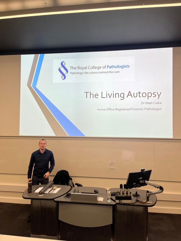 Dr Matt Cieka in front of a presentation screen showing the Living Autopsy cover slide.
