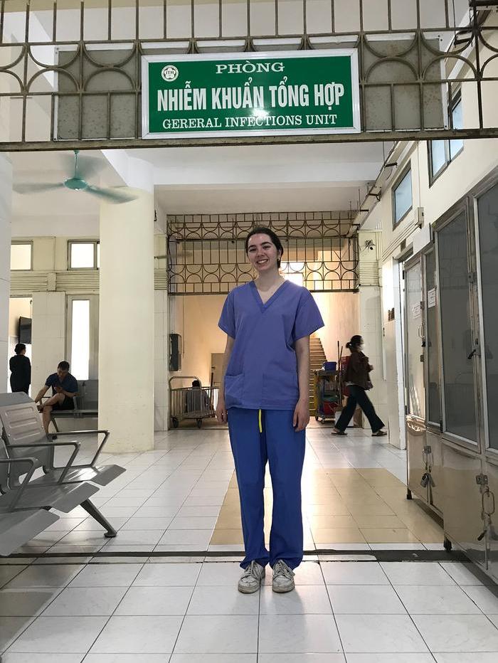 Antonia-Olivia Roberts, in scrubs, smiling, standing under the General Infections Unit sign at Bach Mai Hospital in Hanoi, Vietnam.